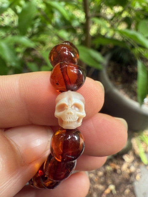 A hand holding a bracelet with translucent amber beads and a carved skull centerpiece.
