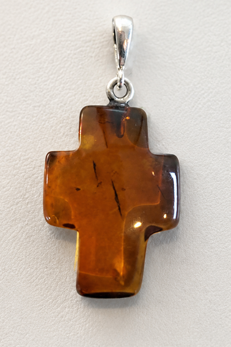 A cross-shaped amber pendant with a polished silver bail, displayed on a light textured background.