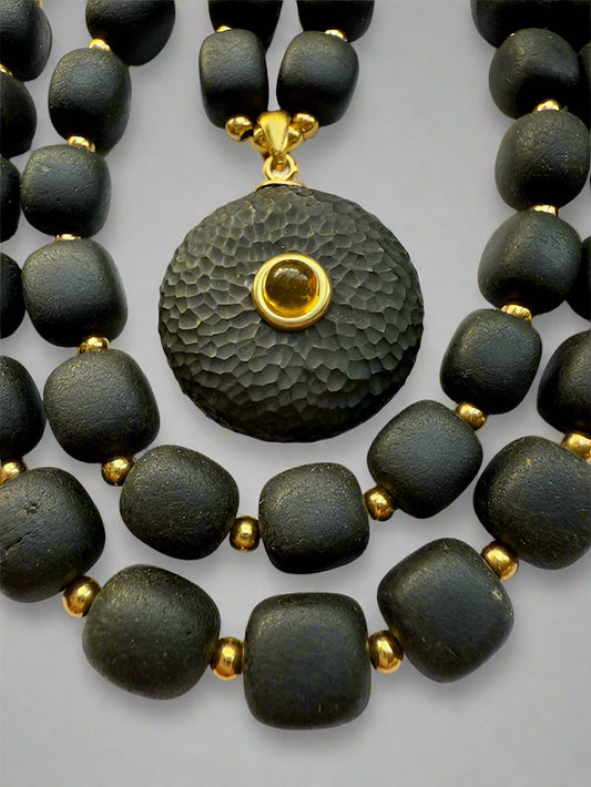 A detailed image of a sculpted amber pendant necklace with dark rounded beads, gold accents, and a polished gold bezel setting.