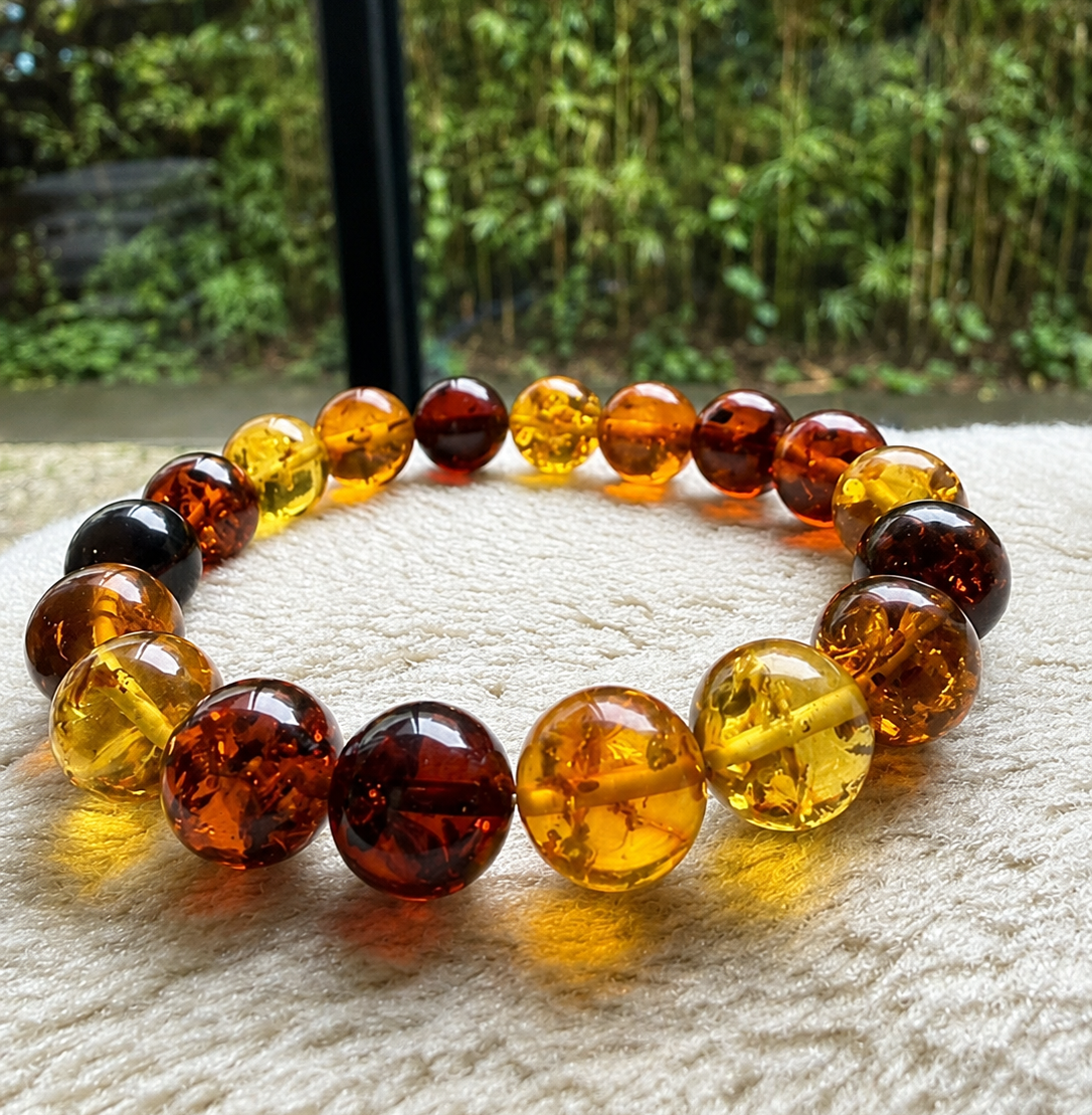 A beaded bracelet made of translucent round amber-like beads in shades of yellow, orange, and dark reddish brown, displayed on a textured cream fabric with a blurred green outdoor background.