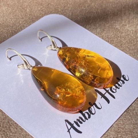 A pair of amber drop earrings laid on a white card with the text 'Amber House' printed below.