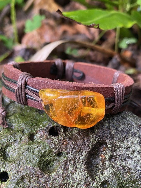 A brown leather bracelet with a large cognac amber stone clasp, displayed on a natural outdoor background.