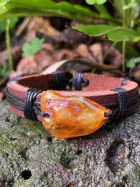 A brown leather bracelet with a large, raw amber stone set in a black leather loop, displayed on a natural outdoor background.