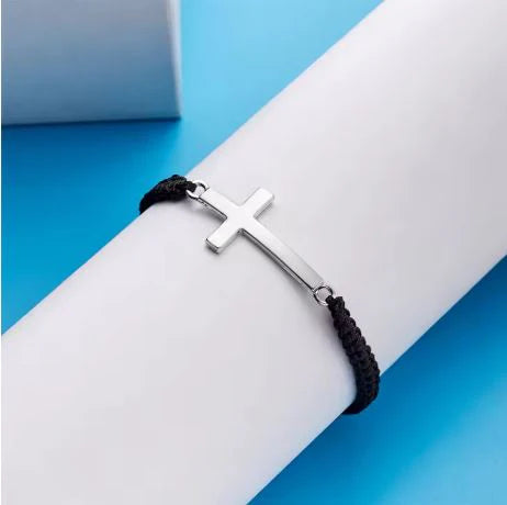 Silver cross bracelet on a black cord, displayed on a white cylindrical object with a blue background.